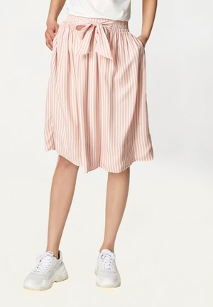Vrouw draagt een knielange roze-wit gestreepte rok met een gestrikte strik in de taille en witte sneakers, handen in de zakken, staand op een witte achtergrond.