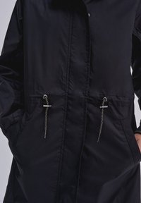 Černá voděodolná bunda se zipem vpředu, dvěma bočními kapsami a nastavovacími šňůrkami v pase. Hladký, lesklý materiál.