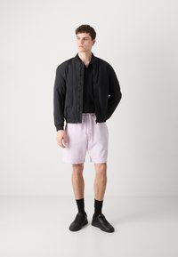 Sort bomberjakke med en glat overflade, parret med lyserøde shorts lavet af blødt stof. Sorte sneakers fuldender outfittet.