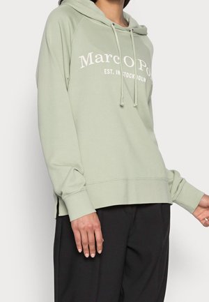 Person trägt hellgrünen Hoodie mit dem Schriftzug „Marco Polo“ und schwarze Hose, steht vor einem schlichten hellen Hintergrund.