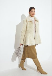 Faux fur cremefarvet frakke, lang beige ruskindsnederdel, knæhøje beige støvler med spidse tæer og en fransedekoreret lys grå håndtaske.