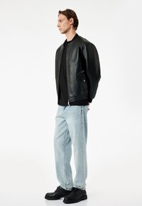 Chaqueta bomber de cuero negro con puños acanalados, combinada con jeans de mezclilla azul claro y zapatos negros. El modelo está de lado contra un fondo blanco.