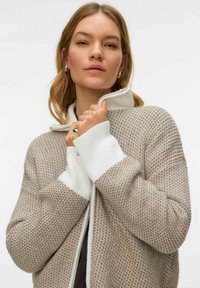 Beež tekstuuriga lukuga kardigan, millel on valged soonikkoes mansetid ja krae, geomeetrilise mustriga ning lõdva lõikega disain.