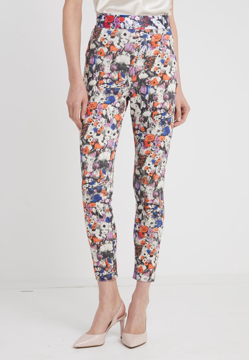 Pantalon slim longueur cheville avec un motif floral multicolore en blanc, orange, violet et bleu, porté avec des chaussures à talons hauts couleur nude.
