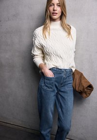 Krémový pletený svetr s texturovaným diamantovým vzorem, doplněný vysokým modrým denimovým kalhotami s předními kapsami. Hnědý textilní doplněk.