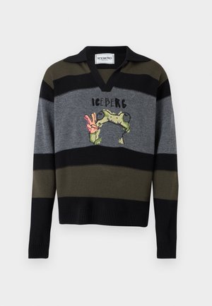 Mand iført solbriller, stribet sweater med frø-grafik og teksten "Iceberg", brede brune cargobukser og røde sneakers, stående med hånden i lommen.