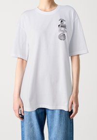 T-shirt blanc oversized avec un petit graphique noir et un texte japonais sur le coin supérieur gauche de la poitrine, porté avec un jean en denim bleu clair.