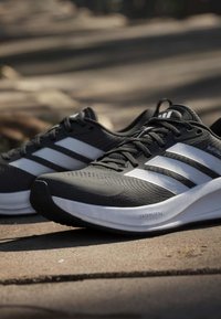 Chaussures de course Adidas noires et blanches avec un design à lacets reposant à l'extérieur sur une surface pavée à la lumière naturelle.