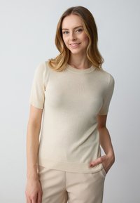 Maglione a maniche corte in beige, con scollatura tonda e orlo a costine. Texture liscia, forma aderente. Abbinato a pantaloni color crema.