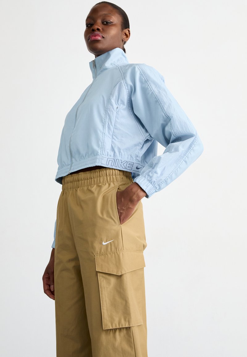 Giacca cropped blu chiaro con tessuto testurizzato e chiusura con zip; abbinata a pantaloni cargo beige con tasche e un piccolo logo bianco.