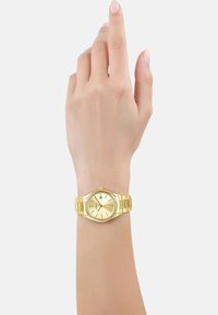 Gold stainless steel watch with a round face, simple dial, and date feature, worn on a hand with a neutral skin tone.