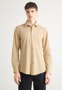 Camisa beige de manga larga, con botones en el frente y un patrón texturizado, y cuello extendido. Combinada con pantalones oscuros.