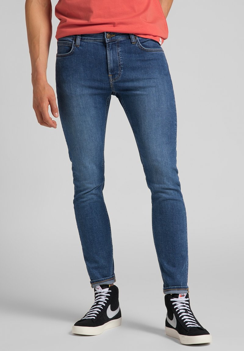 Jeans de denim azul ajustados con un ligero estiramiento, diseño clásico de cinco bolsillos y puños enrollados, combinados con zapatillas altas negras.