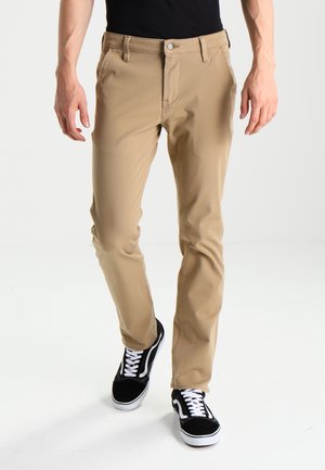 Spodnie khaki o wąskim kroju, zapięcie na guzik, szlufki na pasek i subtelne przednie kieszenie, w zestawieniu z czarnymi trampkami z białymi sznurówkami.