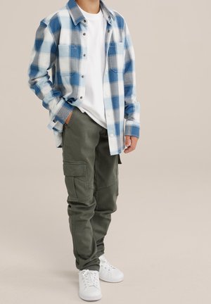 Barn iført grønne cargobukser, hvide sneakers, hvid T-shirt og en blå- og hvidternet knap-op skjorte, står med den ene hånd i lommen.