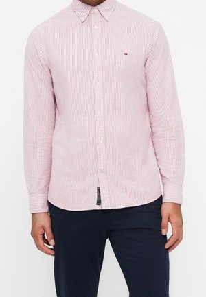 Hombre con una camisa de rayas rosas y blancas de manga larga abotonada, con pantalones oscuros, de pie con las manos relajadas a los lados.