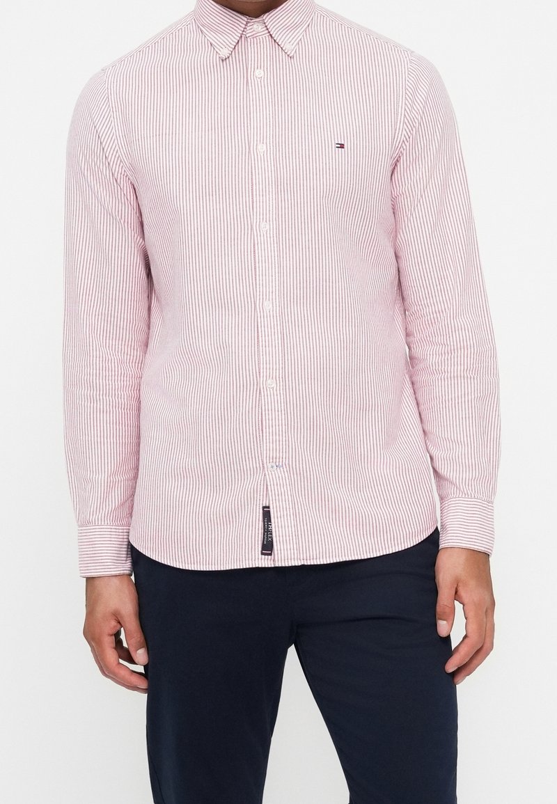 Homme portant une chemise rayée rose et blanche à manches longues, boutonnée, avec un pantalon foncé, debout, les mains détendues le long du corps.