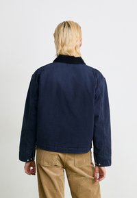 Person with short blonde hair wearing a navy jacket with black collar and tan corduroy pants, facing away against plain background.