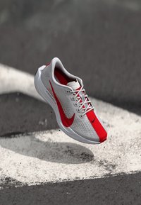 Chaussure de course légère avec une tige en mesh gris, accents rouges et semelle blanche. Présente des perforations et des lacets plats pour un ajustement sécurisé.