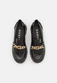 Mocassins en cuir verni noir avec une grosse chaîne dorée en accent et une semelle en caoutchouc. Caractérisés par un bout arrondi et des détails de coutures texturées.