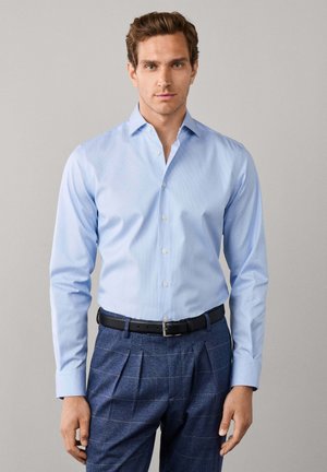 Man draagt een lichtblauw overhemd met lange mouwen en knopen en donkerblauwe geruite broek met een zwarte riem, staand voor een effen grijze achtergrond.