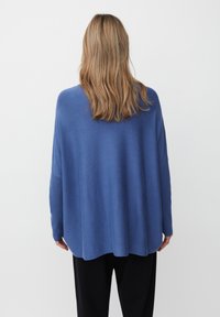 Person mit schulterlangen, hellbraunen Haaren, der einen lockeren blauen Pullover und schwarze Hosen trägt und vor einem schlichten weißen Hintergrund steht.