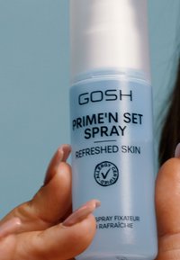Blauwe plastic sprayfles met een witte dop, gelabeld "GOSH PRIME'N SET SPRAY REFRESHED SKIN," inclusief een allergiecertificeringsicoon.