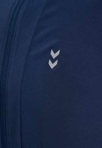Marineblåt jakke stof med en lodret lynlås til venstre og to lysegrå nedadvendte Chevron-logoer på højre side.