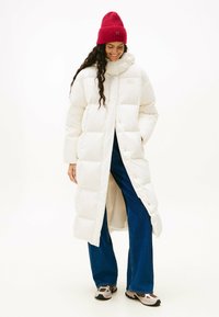 Witte pufferjas met een capuchon, voorzien van vierkante gewatteerde secties. Gepaarde met donkerblauwe wijde jeans en een rode geribbelde beanie.