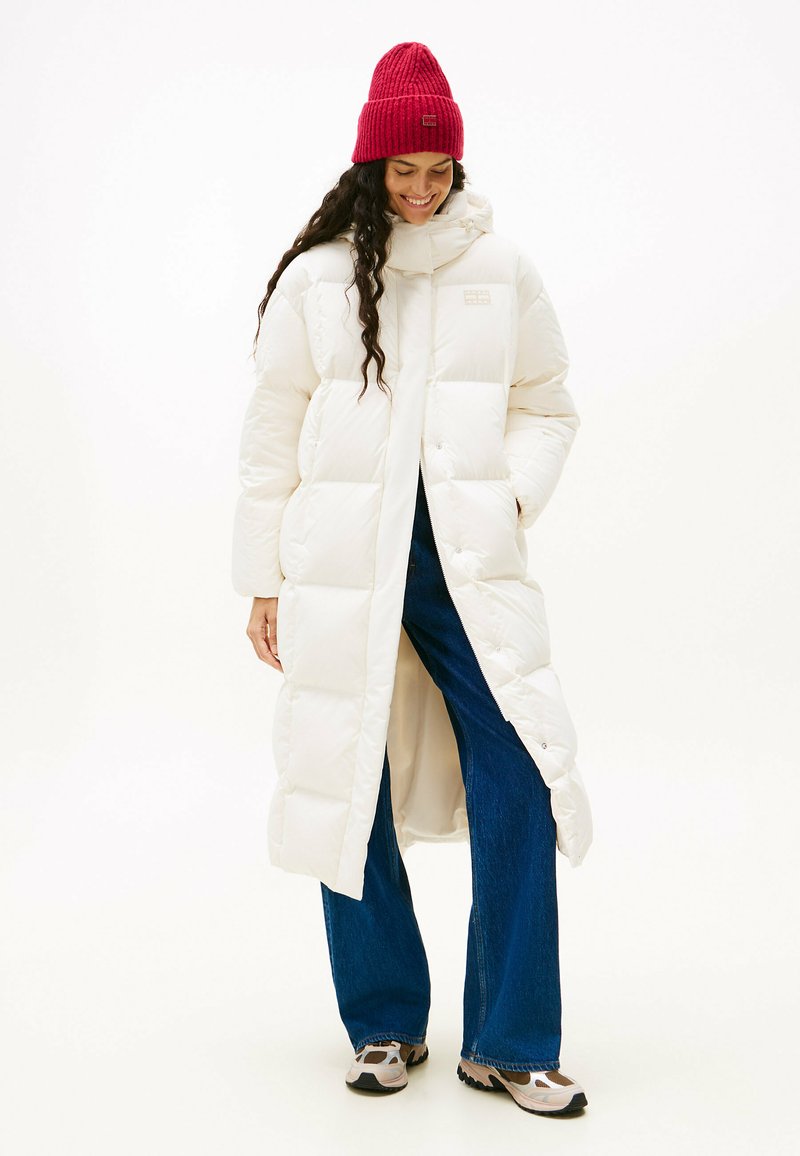 Witte pufferjas met een capuchon, voorzien van vierkante gewatteerde secties. Gepaarde met donkerblauwe wijde jeans en een rode geribbelde beanie.