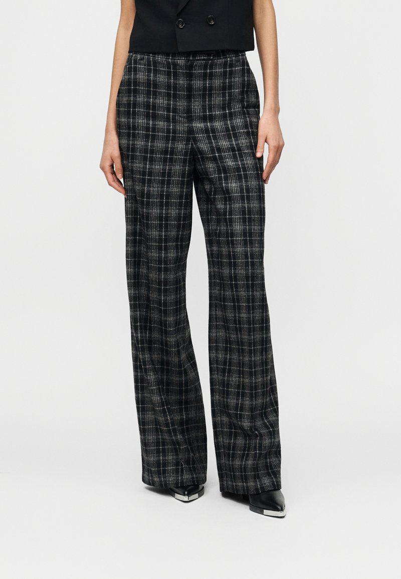 Γυναίκα που φοράει φαρδιά καρό παντελόνια σε μαύρο και γκρι χρώμα, με μαύρα μυτερά παπούτσια και αμάνικο μαύρο τοπ, μπροστά σε λευκό φόντο.