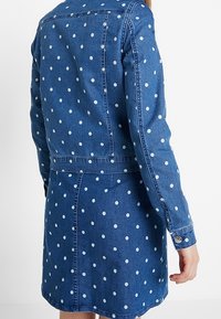 Woman wearing a blue denim jacket and skirt set with white polka dots, shown from the back with her right hand relaxed by her side.