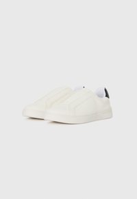 Zapatillas deportivas blancas sin cordones con suelas gruesas y lengüetas de talón negras, mostradas en un ángulo sobre un fondo blanco liso.