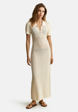 Vrouw draagt een crème-gekleurde, gebreide maxi-jurk met korte mouwen, een kraag en knopen, gecombineerd met bruine platte sandalen.