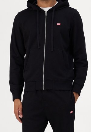Mann trägt einen schwarzen Kapuzenpullover mit Reißverschluss und passende schwarze Hose, beide mit kleinen roten Diesel-Logos, steht mit einer Hand in der Tasche.