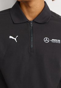 Polo noir avec col zippé, arborant un logo Puma blanc et le branding de l'équipe Mercedes AMG Petronas F1 sur la poitrine. Coupe classique.