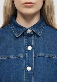 Personne blonde portant une veste en jean bleue avec des boutons argentés, montrant le col et le haut de la poitrine, lèvres fermées et expression neutre.