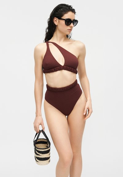 Femme portant un bikini marron asymétrique avec des découpes et des lunettes de soleil, tenant un petit panier tissé noir et beige à rayures.