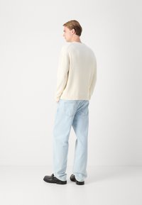 Jersey de canalé crema con mangas largas, combinado con jeans de corte relajado de color azul claro y zapatos negros, visto desde la parte de atrás.