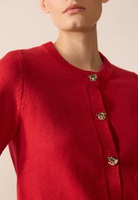Cardigan rouge en tissu tricoté doux, avec un col rond et des accents de boutons métalliques avec une finition texturée le long de la patte de boutonnage.