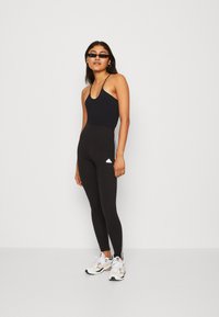 Top preto sem mangas com alças finas, combinado com leggings pretos de cintura alta com riscas laterais brancas e um logótipo. Ténis brancos com detalhes.