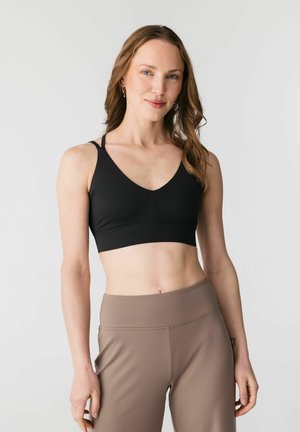 Femeie cu păr ondulat castaniu deschis, purtând un sutien sport negru și colanți taupe cu talie înaltă, stând în picioare pe un fundal simplu și luminos.