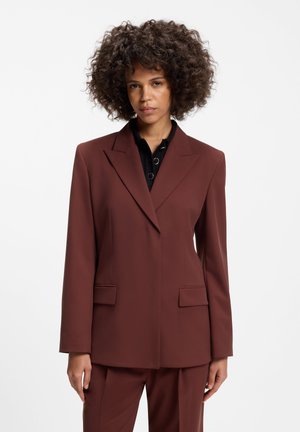 Femme aux cheveux bouclés portant un blazer marron tailleur et un pantalon assorti sur une chemise noire sur fond blanc.