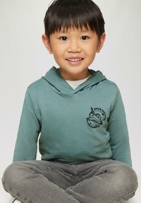 Licht teal hoodie met een buidelzak en een zwart "Space Explorer" grafiek. Grijze jeans, zittende pose met gekruiste benen, neutrale achtergrond.