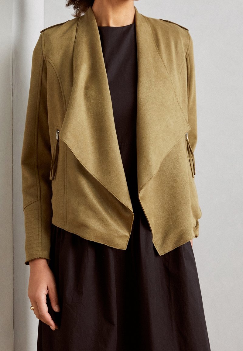 Femme portant une veste drapée vert olive ouverte sur une robe noire, debout devant un mur clair uni.