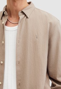 Camisa beige de botones hecha de una tela texturizada con un sutil patrón de espiga, que presenta un pequeño logotipo bordado en el pecho.