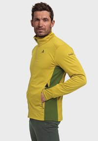 Gelbe und grüne Softshell-Jacke mit hohem Kragen, Reißverschluss vorne und Seitentaschen. Texturiert, figurbetontes Design, mit Logo der Marke am Ärmel.