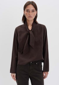 Donna con capelli castani lisci indossa una blusa a maniche lunghe marrone scuro con cravatta e pantaloni scuri, in piedi contro uno sfondo chiaro e semplice.