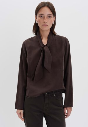 Femme avec des cheveux bruns raides portant un blouse à manches longues marron foncé avec un nœud cou et un pantalon sombre, se tenant contre un fond clair.