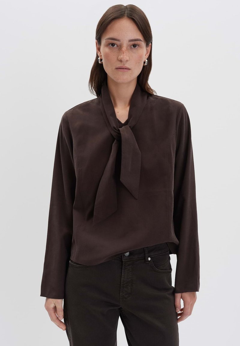 Donna con capelli castani lisci indossa una blusa a maniche lunghe marrone scuro con cravatta e pantaloni scuri, in piedi contro uno sfondo chiaro e semplice.